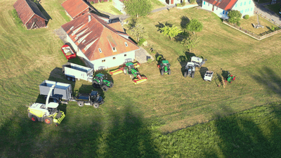 Mehrere landwirtschaftliche Fahrzeuge stehen vor einem Bauernhof auf einem Feld in der Sonne.
